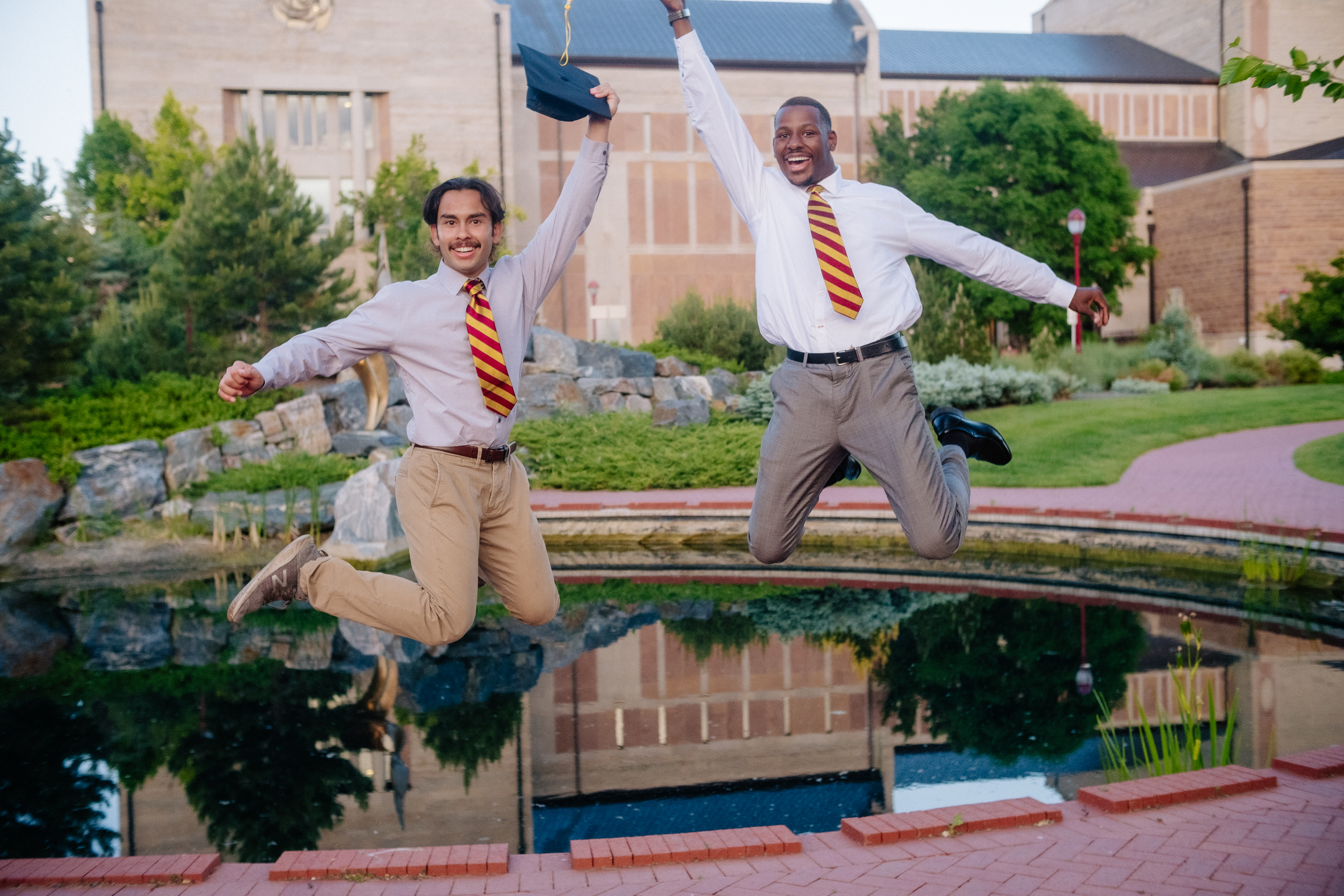 Two graduates jumping for joy by a campus pond at golden hour